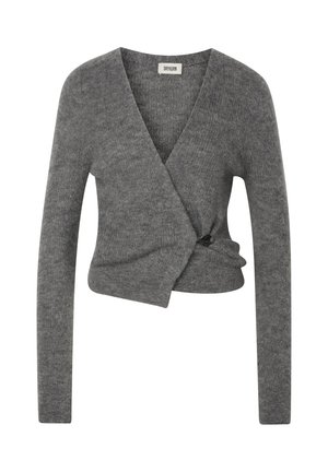 Femme debout les mains dans les poches, portant un pull cache-cœur gris, un jean large avec revers et des bottines noires à talons.