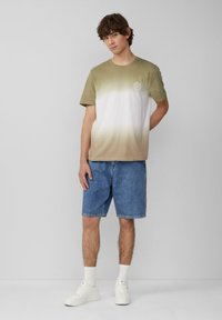 Kurzarm-T-Shirt mit einem Verlauf von olivgrün nach weiß, mit einem kleinen Logo auf der Brust, kombiniert mit Jeansshorts und weißen Sneakers.