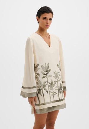 Femme portant une robe longue crème à manches longues avec des imprimés de palmiers gris et un col en V, debout devant un fond clair uni.