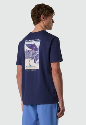 Uomo che indossa una maglietta blu scuro con grafica della spiaggia "French Riviera" e pantaloncini azzurri, in piedi davanti a uno sfondo neutro, di profilo.