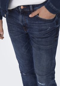 Jeans din denim albastru închis, cu croială slim, având o ușoară uzură, cusături în contrast și un nasture din cupru la talie.