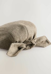Houndstooth-mönstrad hatt i en ljusbrun nyans, med en rund krona och en knuten rosettdetalj bak, tillverkad av texturerad tyg.
