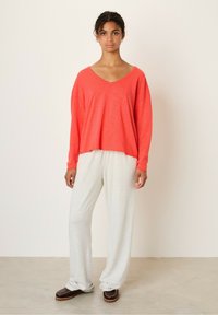 Koralfarvet V-hals sweater med lange ærmer, parret med lysegrå, løstsiddende sweatpants og mørkebrune slip-on sko. Glat stofstruktur.