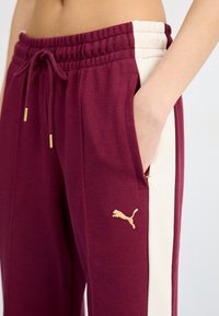 Bordeaux joggers med cremefarvede sidelapper, der har en struktureret stof og gul snøre. Puma-logoet er fremhævet i guld på låret.