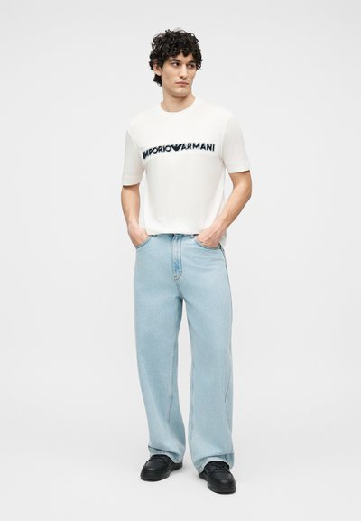 Άνδρας με σκουρόχρωμα σγουρά μαλλιά, φορώντας λευκό t-shirt Emporio Armani, ανοιχτό μπλε τζιν παντελόνι με φαρδιά μπατζάκια και μαύρα παπούτσια, στέκεται με τα χέρια στις τσέπες.