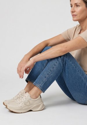Kvinde sidder på gulvet med bøjede ben iført blå jeans, beige T-shirt og beige sneakers med hjerte-design på siden.