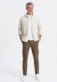 Beige košile s dlouhým rukávem a detaily na knoflíky, pod ní bílá tričko, hnědé cargo kalhoty a bílé tenisky, model stojí na jednobarevném pozadí.
