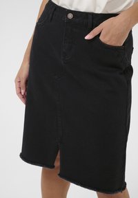 Jupe en denim noir avec une coupe standard, ourlet effiloché, poches avant et une fente subtile en bas. Dotée d'une fermeture à bouton et d'un design classique.