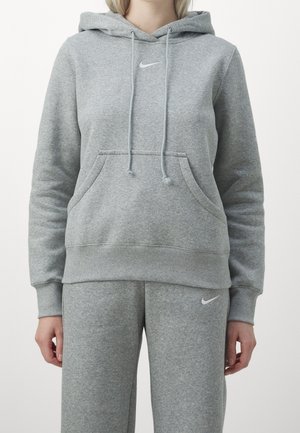 Frau trägt einen grauen Nike-Hoodie mit Kordelzug und passende graue Jogginghose mit Nike-Logos auf der Brust und dem Oberschenkel.