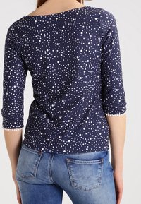 Dunkelbluse mit dreiviertelÄrmeln und einem weißen Punkte-Muster. Der Saum hat einen Spitzenbesatz. Kombiniert mit blauen Jeans.