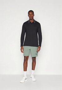 Άνδρας που στέκεται φορώντας μαύρο πουκάμισο με μακριά μανίκια, ανοιχτό πράσινο σορτς με το λογότυπο "BOSS", λευκές κάλτσες και λευκά αθλητικά παπούτσια σε απλή φόντο.