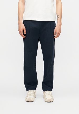 Mężczyzna ubrany w czarne spodnie o prostych nogawkach z fakturą, białą koszulkę i beżowe sneakersy, stojący z ramionami swobodnie opuszczonymi wzdłuż ciała na białym tle.