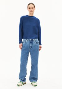 Blauer Pullover mit Rundhalsausschnitt, kombiniert mit hellblauen, lässigen Jeans. Das Outfit wird mit beigen und grünen Sneakern abgerundet.