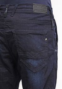 Jeans in denim blu scuro con finitura testurizzata, caratterizzati da tasche posteriori con cuciture decorative e un'etichetta in pelle sulla vita.