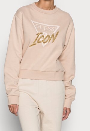 Beżowa bluza z logo trójkąta "GUESS U.S.A" oraz dużym złotym napisem "ICON" o fakturze łańcucha, noszona z dopasowanymi jasnobeżowymi spodniami.