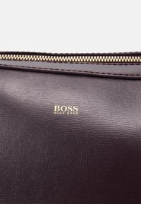 Mørkebrun lædertaske med lynlåslukning og et guldfolieret logo, der står "BOSS HUGO BOSS" på forsiden. Struktureret overflade detalje synlig.