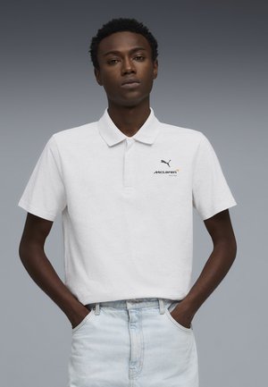 Joven con las manos en los bolsillos, vistiendo un polo blanco de McLaren Racing y jeans azul claro sobre un fondo gris liso.