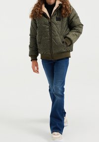 Grön, mönstrad pufferjacka med dragkedja och fleecekrage; kombinerad med blå jeans och ljusa sneakers; har två framfickor.