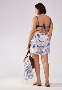 Badetøjsættet har en blå bikinioberdel med justerbare stropper og mønstrede shorts med frugtmotiver. Modellen bærer en strandtaske og et håndklæde.