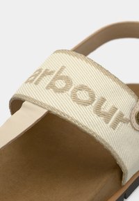 Μπεζ σανδάλι από καμβά με εμφανές λογότυπο, διαθέτει φαρδύ λουράκι, λείο δερμάτινο περίγραμμα και υφή στο πέλμα για άνεση.