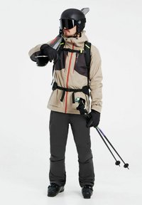 Ski-outfit met een beige en zwarte waterdichte jas met oranje ritsen, zwarte broek, handschoenen, een helm, skibril en skistokken.