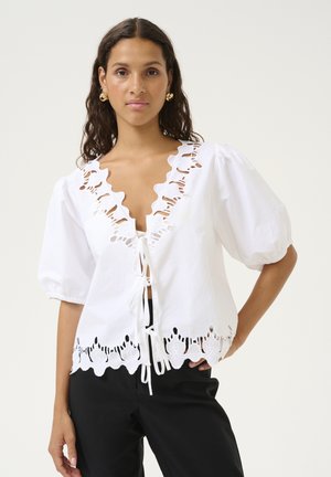 White cotton blouse with a V-neck, ruffled trim, and puff sleeves. Features elaborate cut-out detailing along the hem and a tie closure.
