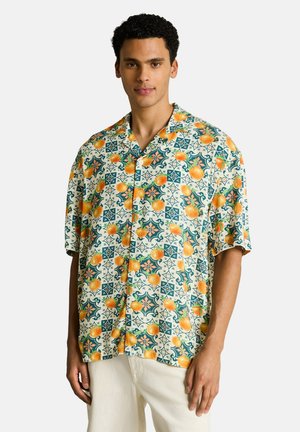 Uomo che indossa una camicia a maniche corte con bottoni, decorata con un motivo di frutta arancione e piastrelle geometriche, abbinata a pantaloni chiari, in piedi davanti a uno sfondo neutro.