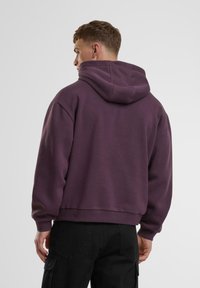 Lila Kapuzenpullover mit großer Kapuze, gerippten Bündchen und Saum, aus weichem Material. Getragen mit schwarzen Hosen, von hinten betrachtet.