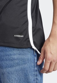 Czarna koszulka sportowa z białymi paskami po bokach. Posiada oznaczenie "AEROREADY" w pobliżu dołu. Noszona z niebieskimi dżinsami, ukazującymi fakturę materiału.