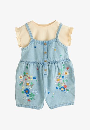 Žuta košulja kratkih rukava s naboranim rukavima u kombinaciji s plavim traper kombinezonom ukrašenim višebojnim cvjetnim vezom i gumbima sprijeda.