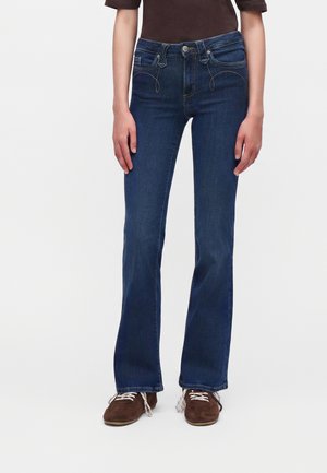 Femme portant un jean bootcut bleu foncé et des chaussures marron à lacets, debout devant un fond uni.