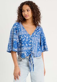 Crop top bleu avec un décolleté en V, des manches courtes en forme de cloche et un design à nouer à l'avant. Décoré de motifs paisley et floraux blancs. Texture en mélange de coton.
