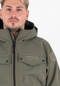 Man met kort blond haar die een olijfgroene jack met ritssluiting draagt, met een "NAPA" patch en een rond embleem op de mouw, recht vooruit kijkt.