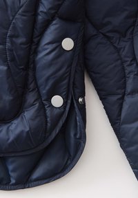 Giacca imbottita trapuntata blu navy con bottoni a pressione argentati e texture liscia, dotata di cuciture curve e un design con imbottitura leggera.