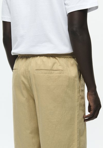 Gros plan d'une personne portant un pantalon beige avec une taille élastique et une chemise blanche à manches courtes, montrant les poches arrière et une main sur le côté.