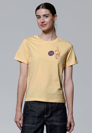 T-shirt en coton jaune avec un col rond et des manches courtes, présentant un design de dessin animé et un texte sur le devant. Porté avec un jean en denim foncé.