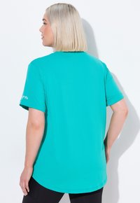 Žena s blond bob účesem, která má na sobě volné tyrkysové tričko s krátkým rukávem a černé legíny, otočená zády k bílému pozadí.