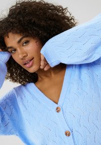 Světle modrý pletený cardigan s výstřihem do V, se vzorem vln a dvěma dřevěnými knoflíky vpředu, měkká textura a volný střih.