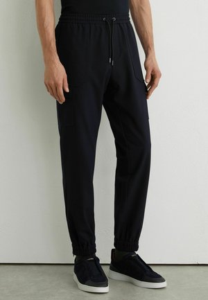 Mand iført sorte joggebukser med sidelommer og sorte sneakers med hvide såler, stående mod en lys væg.