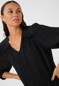 Blouse noire texturée avec un décolleté en V, un détail froncé aux épaules et des manches volumineuses. Le tissu a une apparence froissée.