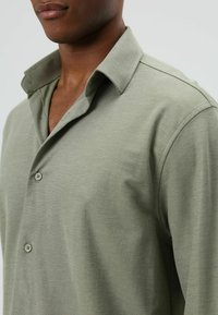 Man draagt een open kraag, lange mouwen, licht olijfgroen overhemd met knopen, waarbij een deel van zijn borst en nek zichtbaar is.