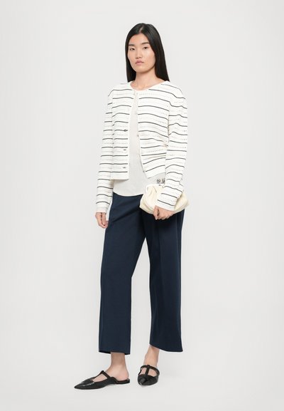 Weiße gestreifte Strickjacke mit LochmusterDetails, kombiniert mit einem cremefarbenen Oberteil, navyblauen weiten Hosen und schwarzen Slippern, während sie eine cremefarbene Clutch hält.