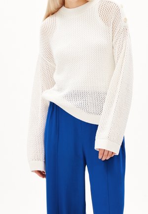 Femme portant un pull en maille blanc avec des boutons sur l'épaule et un pantalon large bleu vif.