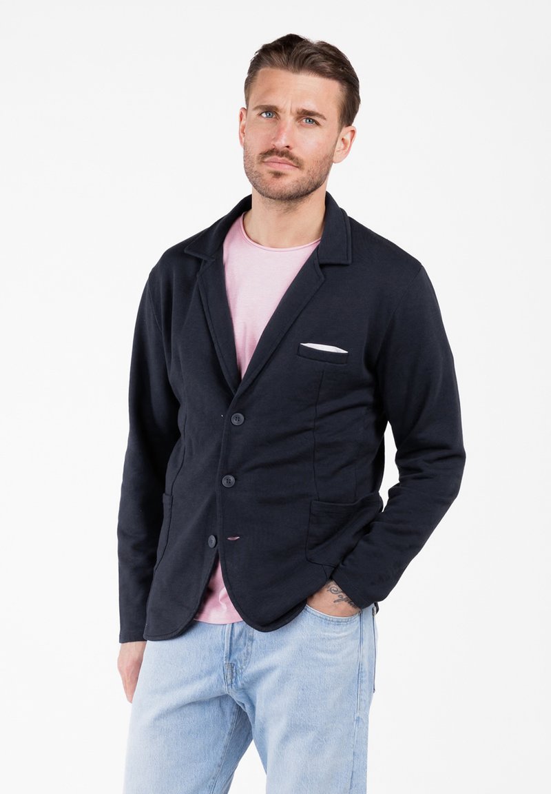 Uomo con capelli corti castani e barba, che indossa un blazer nero su una camicia rosa e jeans azzurri chiari, in piedi con una mano in tasca.