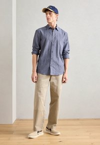 Chemise à boutons bleue à carreaux, pantalons beiges à jambes larges et chaussures beiges à enfiler avec des accents marins ; look décontracté avec les manches retroussées.