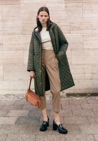 Cappotto lungo imbottito verde con cappuccio, collo alto beige, pantaloni a quadri, scarpe platform nere e borsa a spalla marrone.