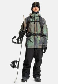 Sněžný snowboardista v maskáčové voděodolné bundě, černých kalhotách na sníh, rukavicích a lyžařských brýlích, drží snowboard a lyžařskou hůl.