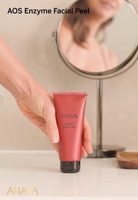 Peeling facial enzimático AHAVA AOS en un tubo rojo mate con tapa negra, de perfil delgado y etiqueta de texto blanco, sostenido por una mano cerca de un espejo.