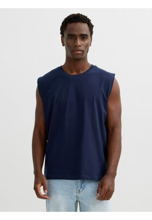 Hombre con camiseta sin mangas azul marino y vaqueros azul claro de pie frente a un fondo blanco liso.