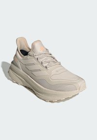 Svetlo béžová športová obuv s textúrovaným syntetickým zvrškom, logom s tromi pruhmi a oranžovými akcentmi. Má hrubú gumovú podrážku a šnurovací dizajn.